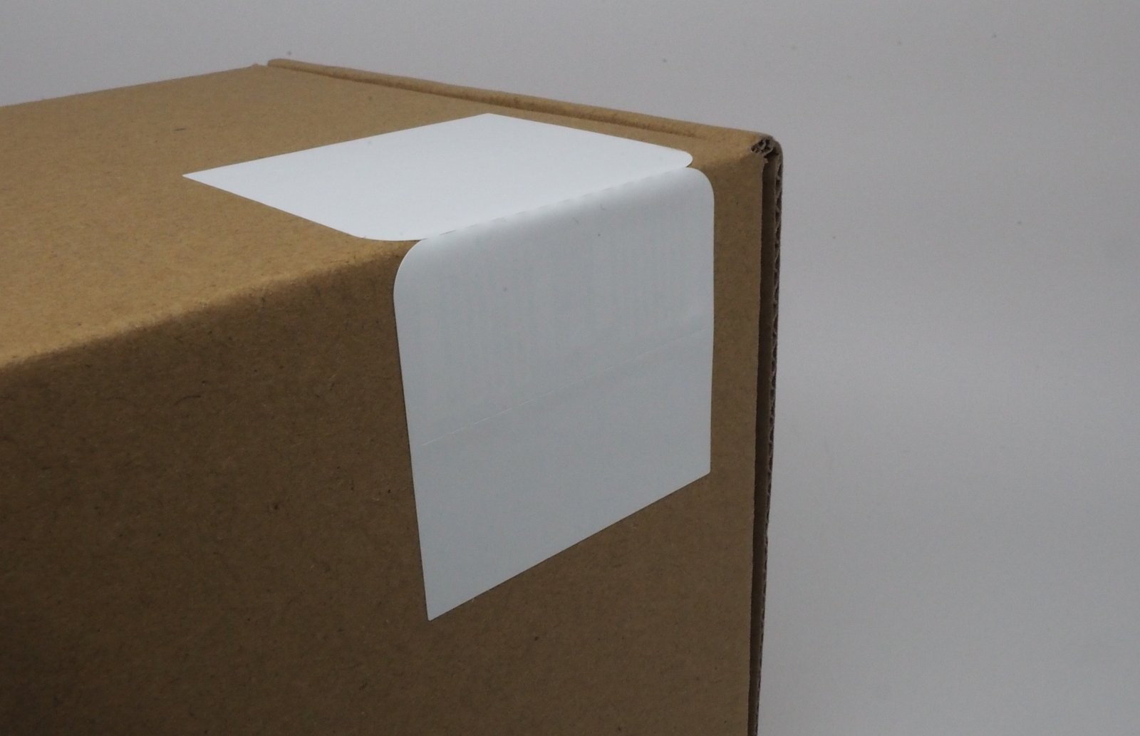 Brown cardboard box close-up with a blank white label on its corner against a plain background. Simple and neutral setting.