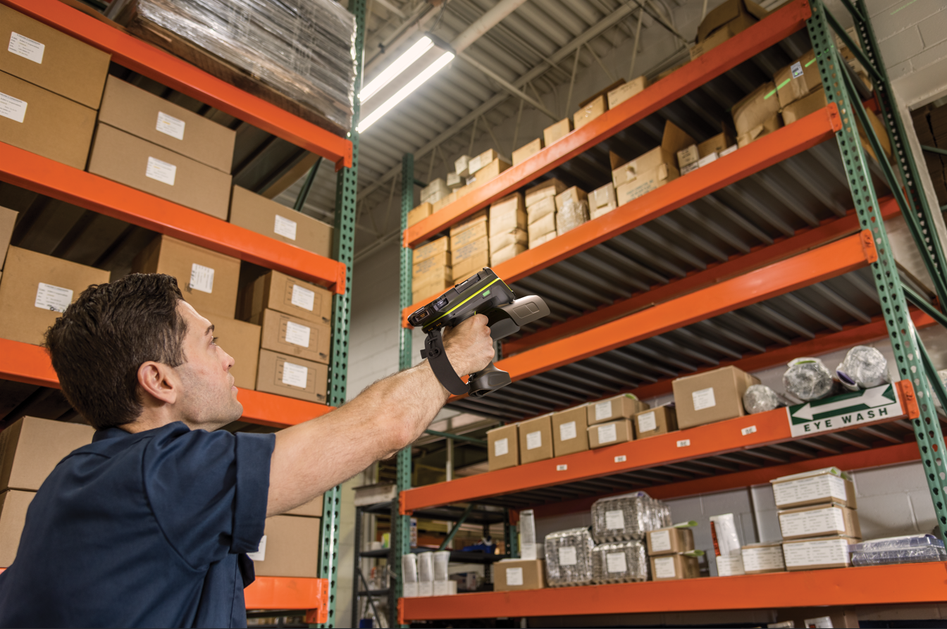 Warehouse worker scans boxes on orange shelves with a handheld device. Cardboard boxes are stacked neatly. Sign reads "Eye Wash."