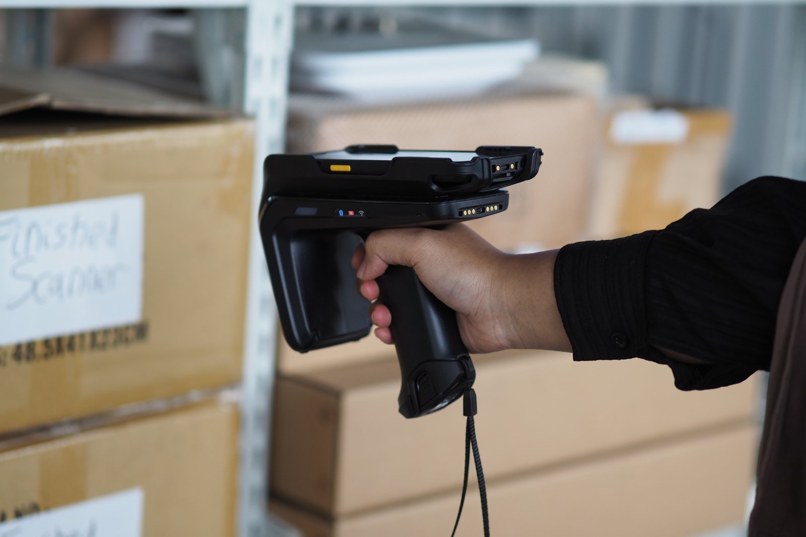 Hand holding a black scanner in a warehouse with labeled boxes in the background. Text on box: "Finished Scanner."