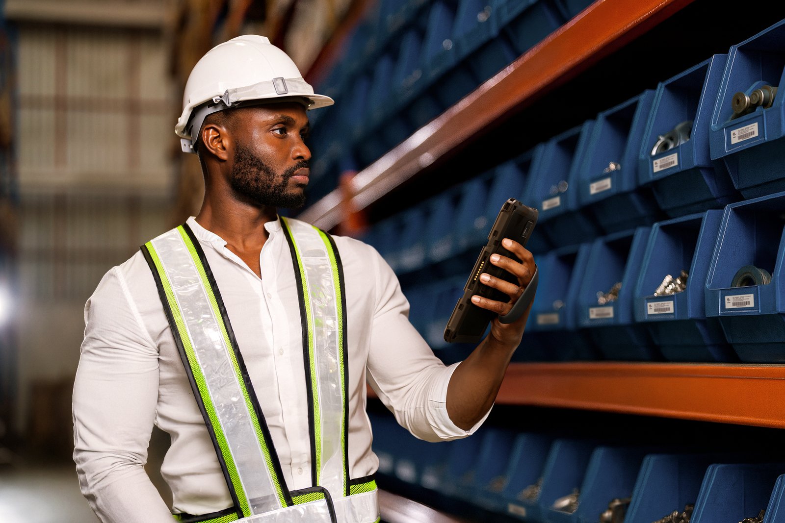 Man in a white shirt and hard hat, holding a tablet, stands in a warehouse with blue bins. He looks focused and wears a safety vest.