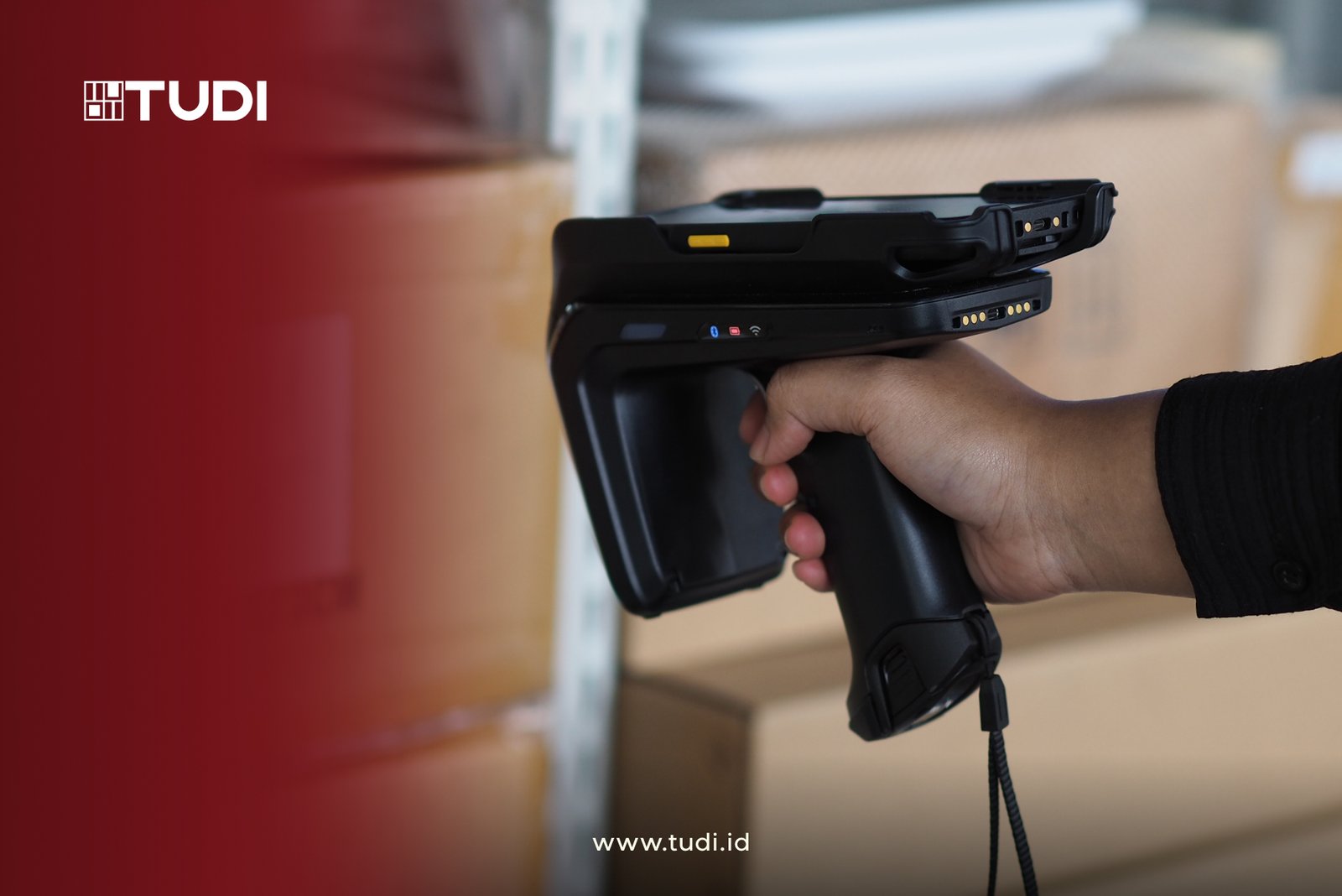 A hand holds a black barcode scanner near brown boxes. Red and white backdrop with text: "TUDI" and "www.tudi.id".
