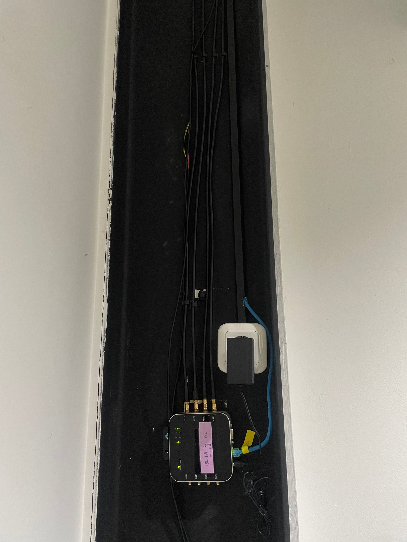 Panel jaringan hitam dengan banyak kabel di ceruk dinding putih, lampu hijau menyala, ada label antena dan catatan ungu.
