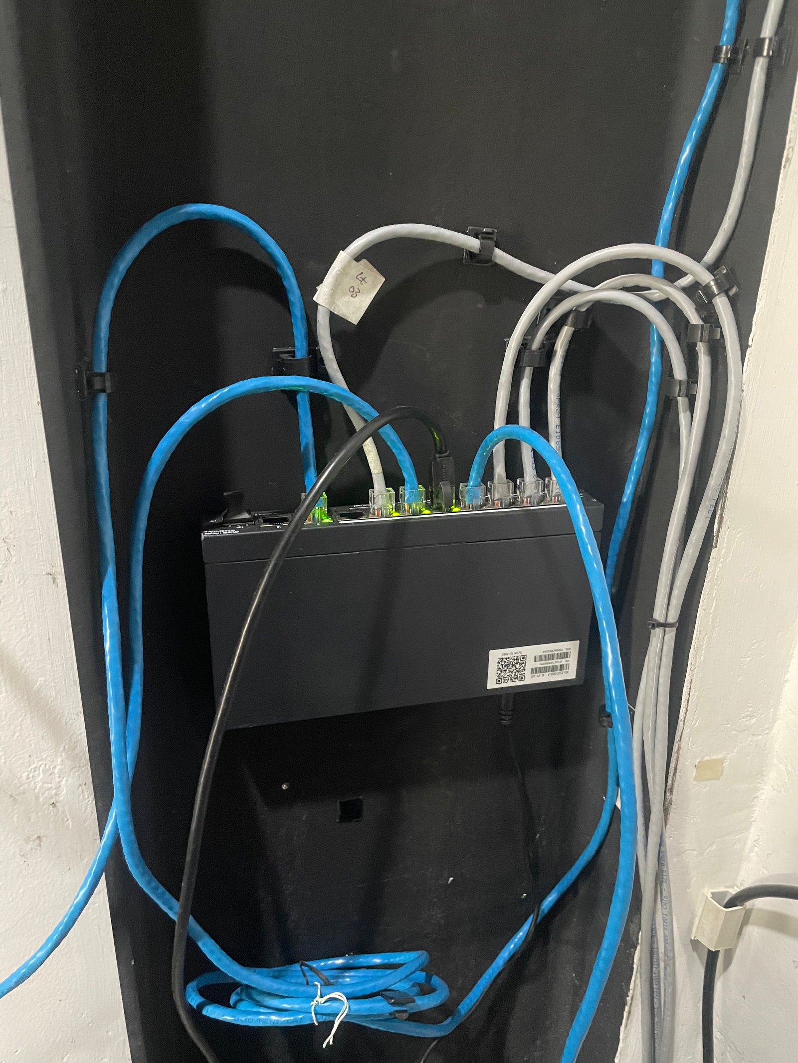 Panel jaringan dengan switch hitam dan kabel biru/abu-abu melingkar, lampu LED hijau menyala; tampak rapi tapi padat.