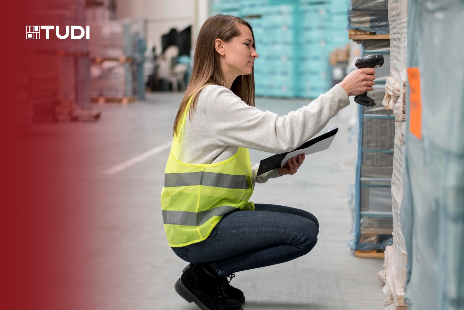A woman wearing a yellow safety vest scans packages in a warehouse using a barcode scanner, holding a street sign. The background shows shelves wrapped in blue plastic.