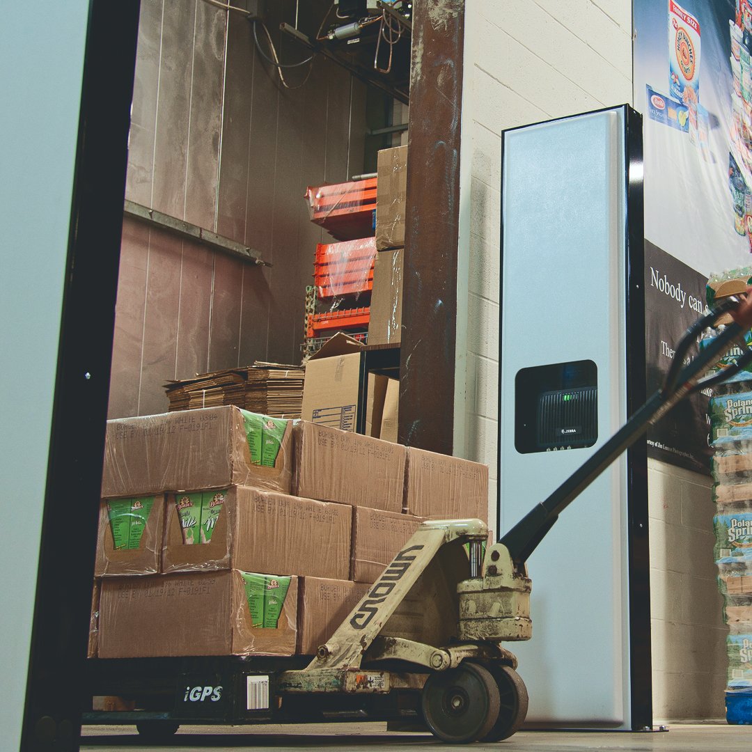 A pallet jack carries brown boxes in a warehouse with stacks of cardboard and posters on the wall. Dim lighting sets an industrial mood.