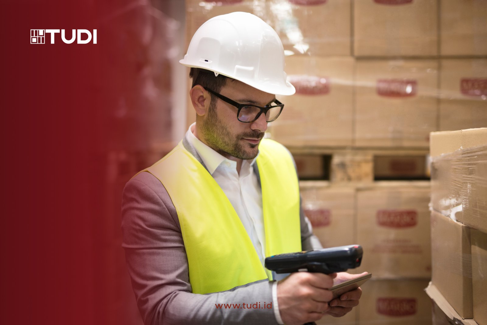 Man in hard hat and yellow vest scans boxes in a warehouse. Background shows stacked cardboard boxes. Logo TUDI, website tudi.id visible.