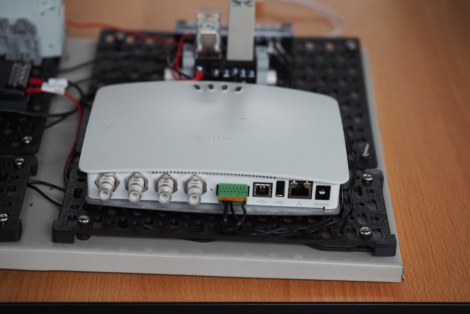 White router with multiple ports on a black mounting board, placed on a wooden surface. Visible text: "ZEBRA" and "24VDC".