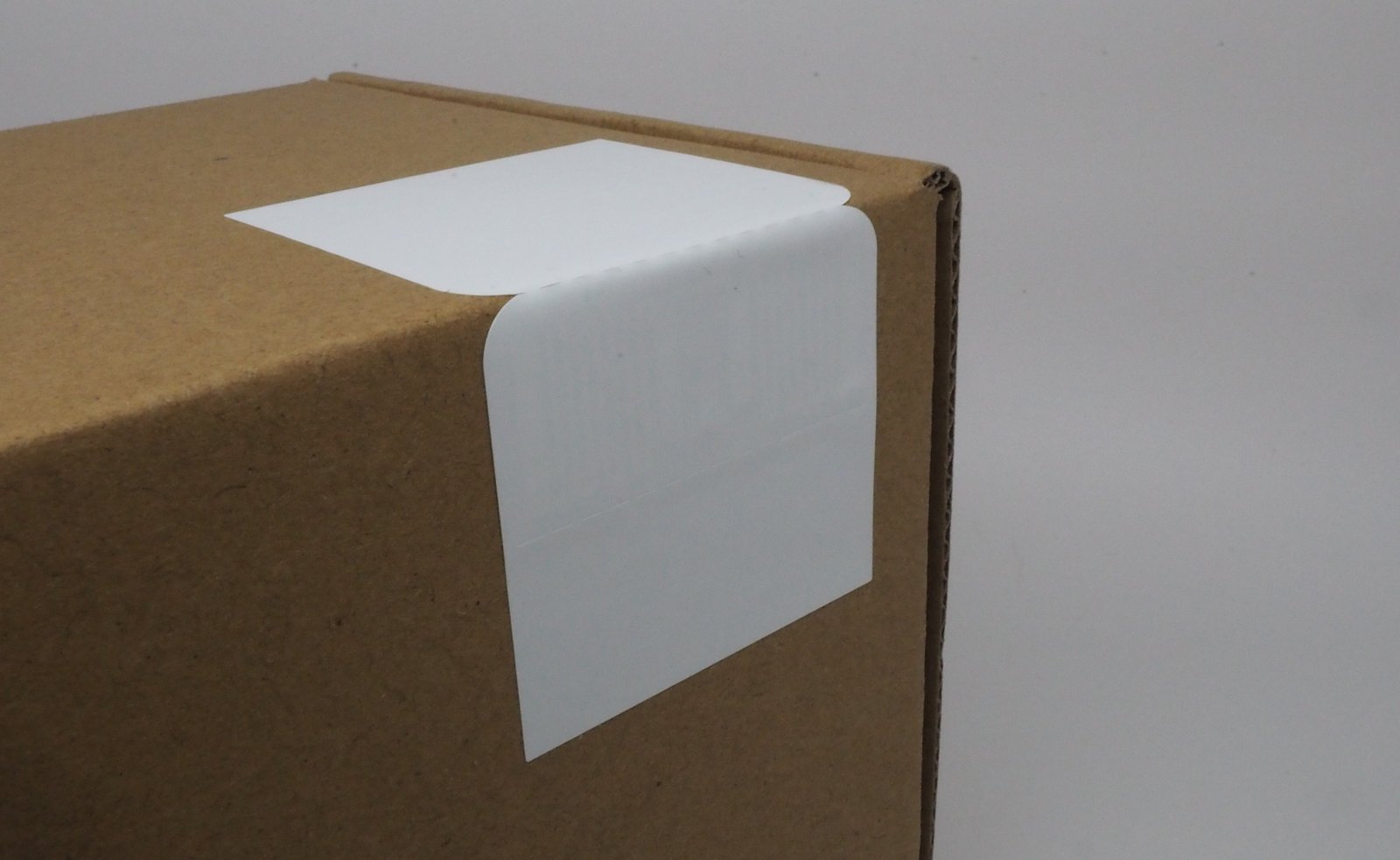 Corner of a brown cardboard box with a white RFID label on the edge. Simple, minimalistic setting with a plain background.
